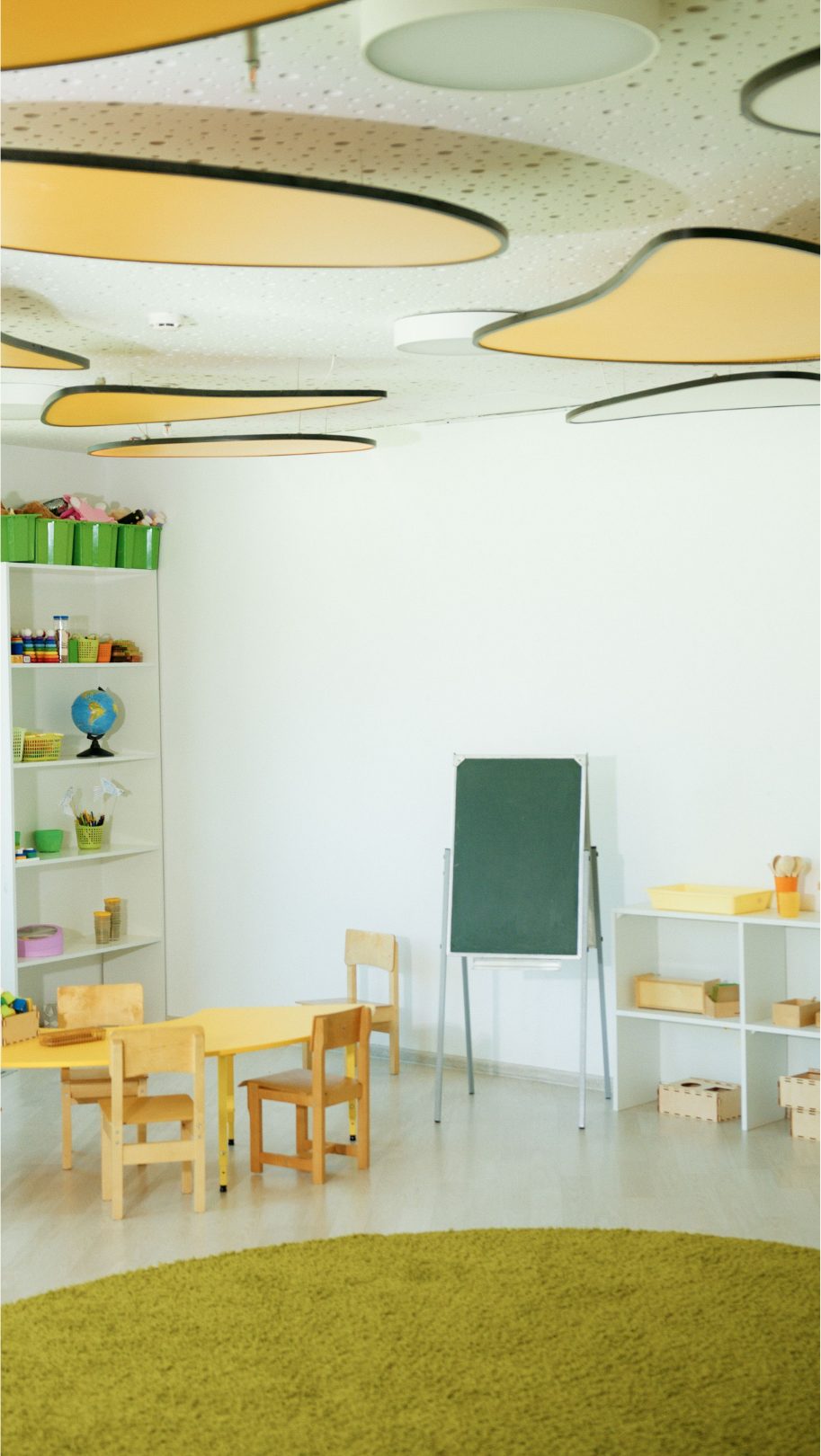 Aula de infantil con mesa amarilla, sillas de madera, pizarra de caballete y estanterías con material